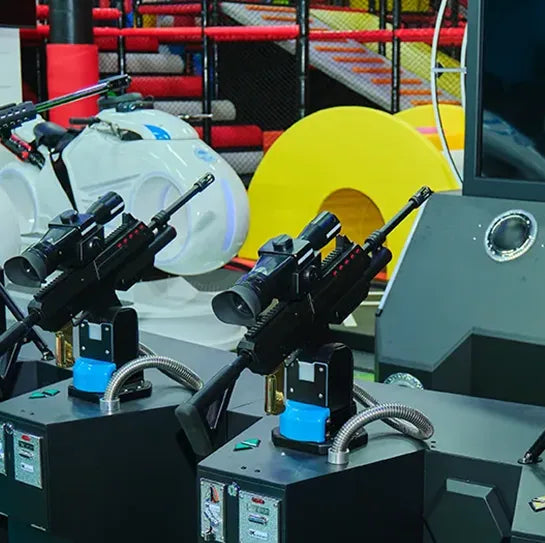 Two arcade-style shooting game machines with attached rifles are in the foreground. In the background, colorful play equipment and slides are visible, suggesting an indoor entertainment or amusement center.