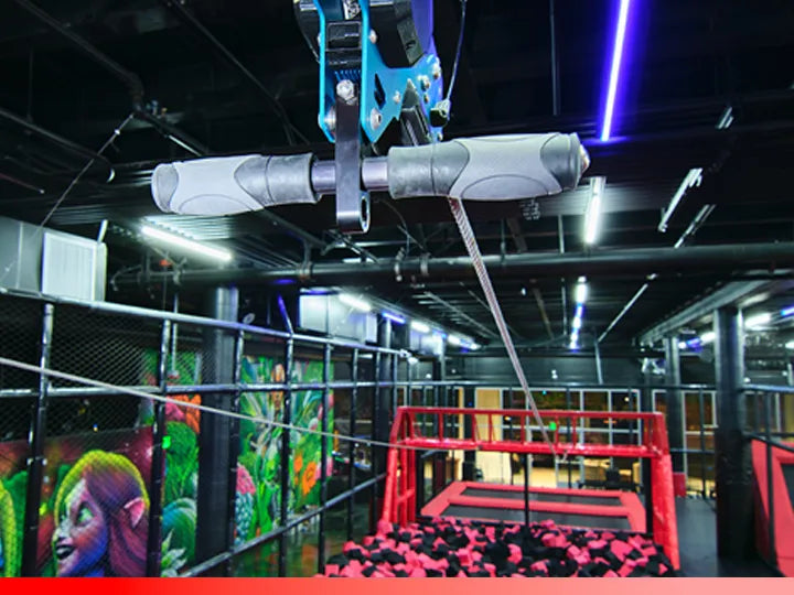 A zipline handle hangs above a foam pit in an indoor adventure park with neon lights, colorful wall art, and safety nets surrounding the area.