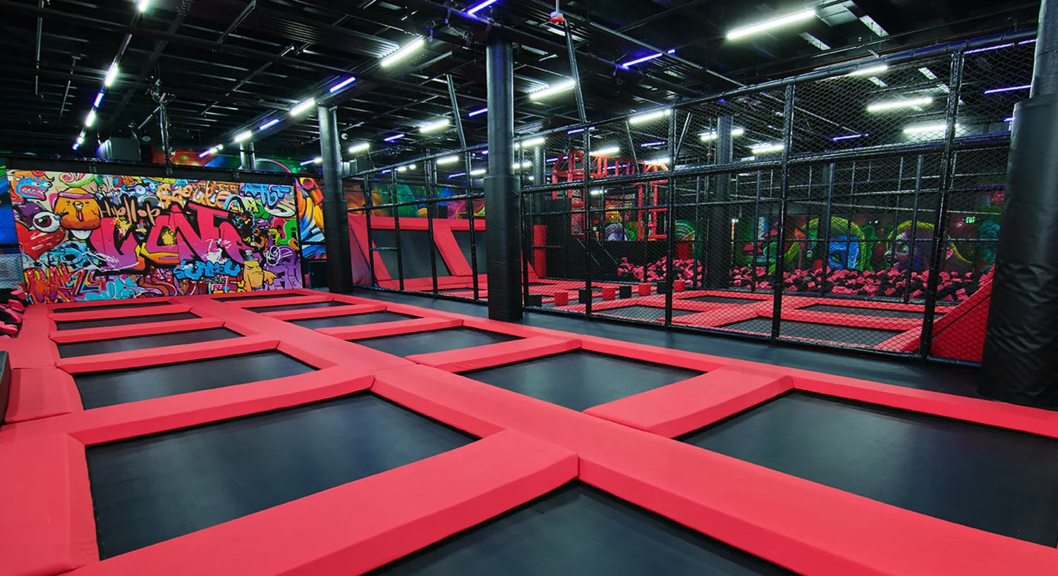 Indoor trampoline park with interconnected red trampolines, black walls, and vibrant graffiti art. The area is well-lit with purple and blue neon lights, and there are safety nets along the sides.