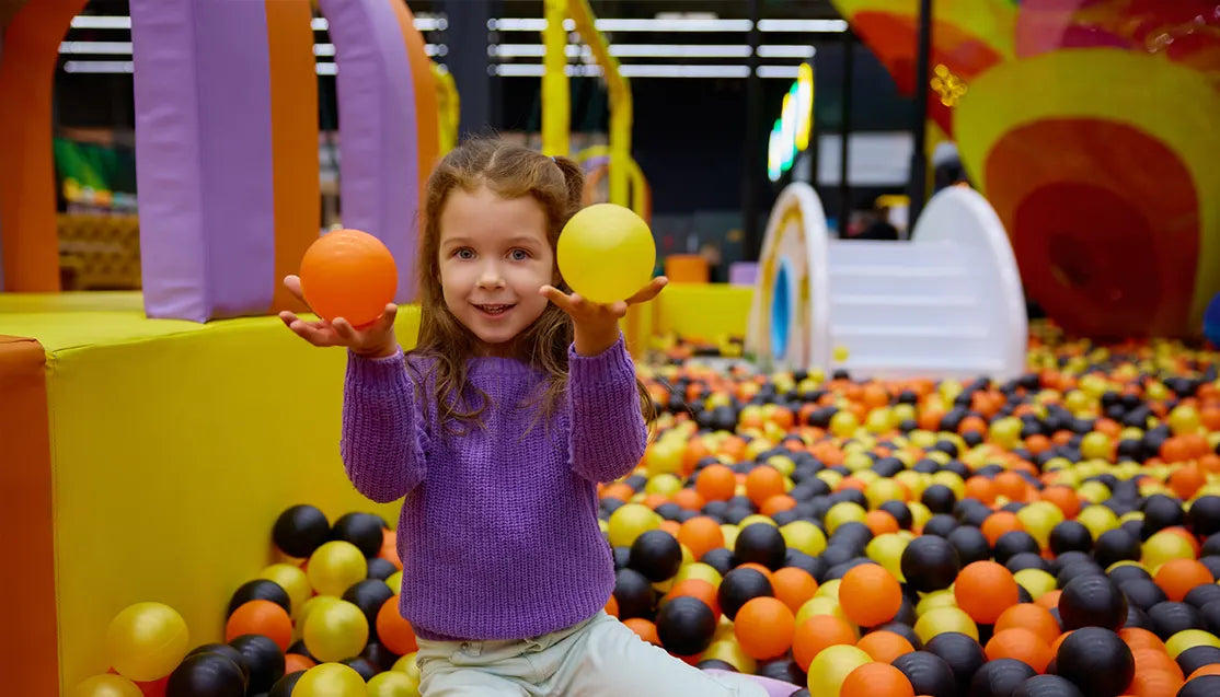 A young girl in a purple sweater sits in a colorful indoor ball pit, smiling and holding up an orange ball and a yellow ball in each hand. The playground is filled with multicolored balls and bright play structures.