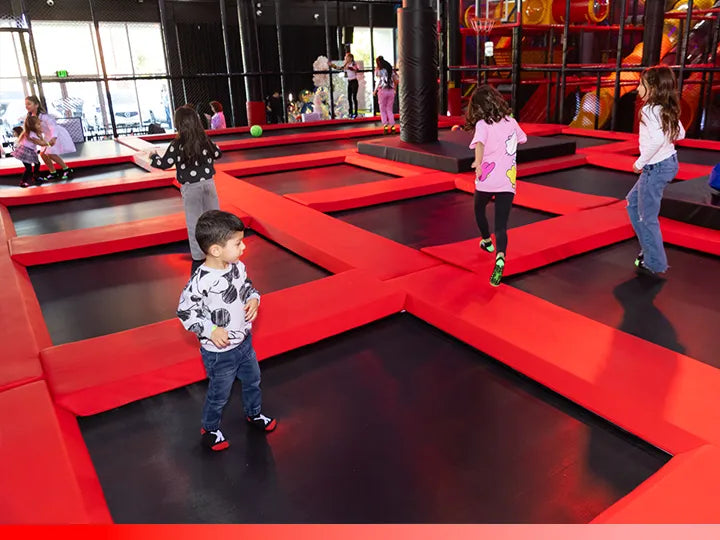 Several children play and jump on interconnected red and black trampolines inside an indoor play center, with more play structures and people visible in the background.